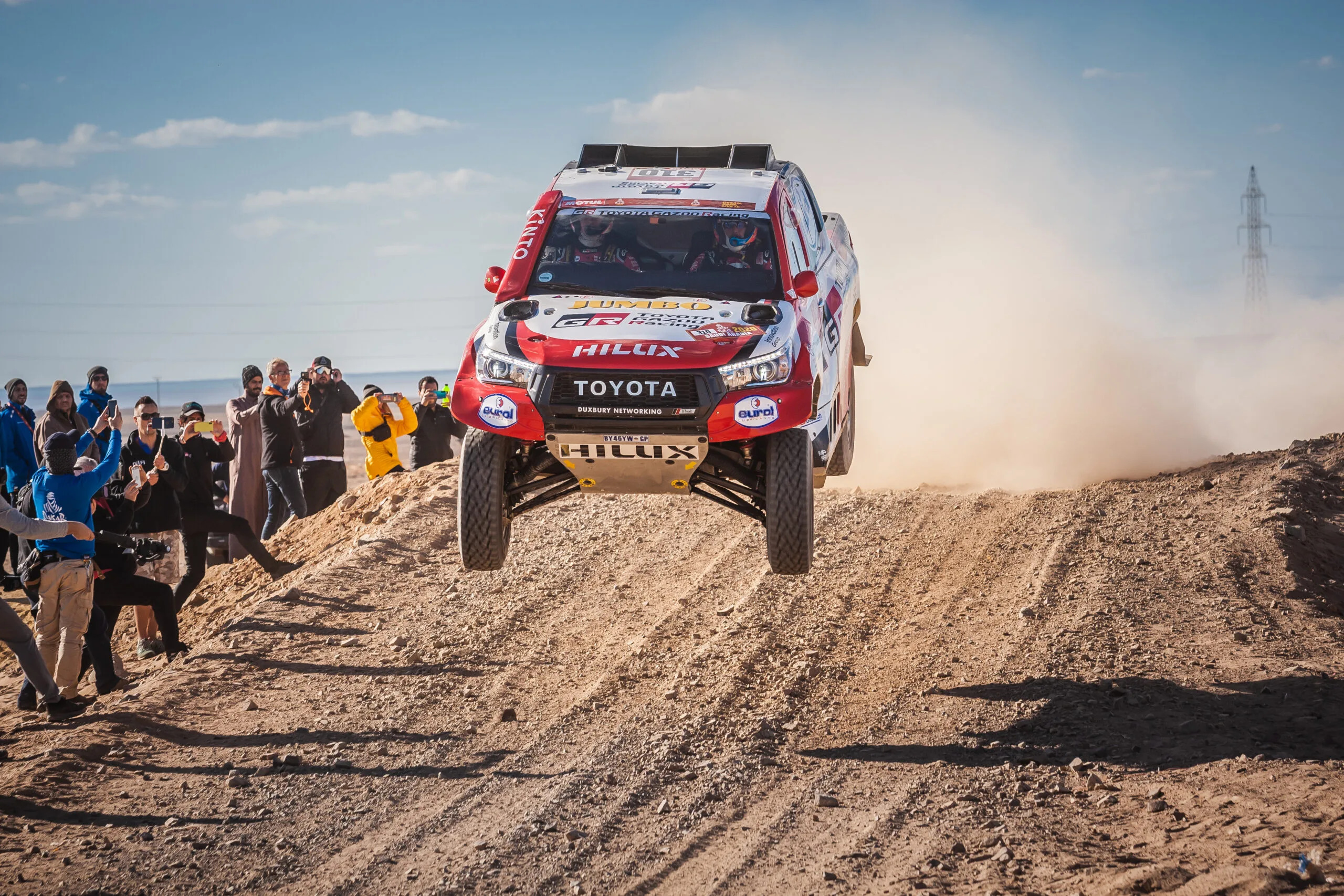 Rally raid car jumping over a gravel road alongside the crowd